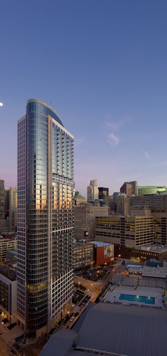 Tall modern skyscraper with reflective glass windows stands among other buildings in an urban cityscape at dusk, with a pool visible on a nearby rooftop.