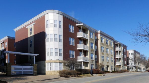 A modern mid-rise apartment building with brick and light-colored accents, balconies, and large windows, situated along a quiet street under a clear blue sky.