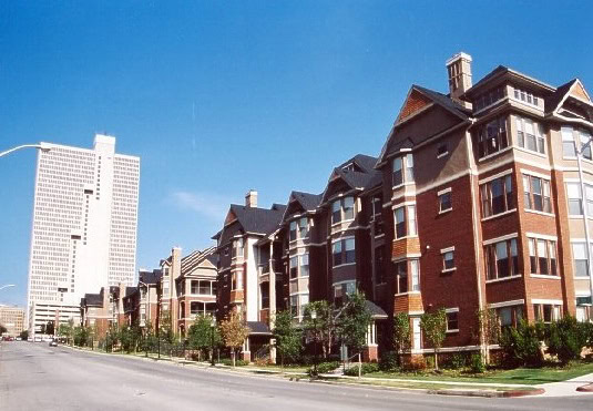 Street view of modern low-rise brick apartment buildings with a tall white high-rise building in the background under a clear blue sky.