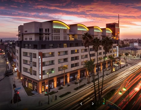 Modern multi-story apartment building on a city corner at sunset, featuring illuminated signage and light trails from passing traffic.