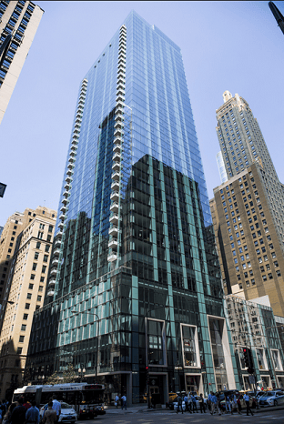Modern glass skyscraper with reflective windows, surrounded by older high-rise buildings and busy street traffic in an urban setting.