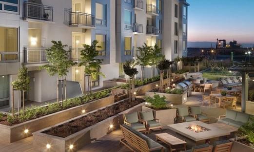 Modern apartment courtyard at dusk with outdoor seating, fire pit, landscaping, and illuminated walkways. Multiple balconies overlook the well-lit communal area.