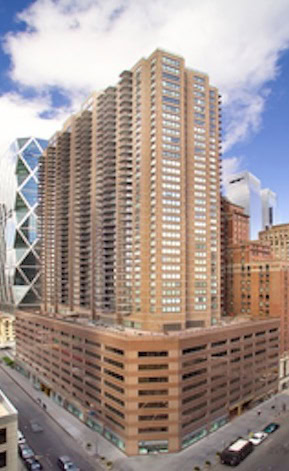 Tall brown high-rise apartment building with many balconies, located on a city corner with cars and pedestrians visible on the surrounding streets.