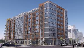 A modern multi-story apartment building with large glass windows and retail space on the ground floor, located at a street corner under a clear blue sky.