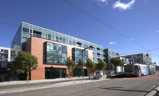 Modern mid-rise apartment building with large windows and a streetcar stopped at a station in front; clear sky and sidewalk with trees visible.