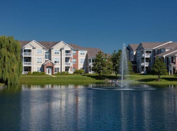Apartment buildings with balconies overlook a pond with a water fountain, surrounded by grass, trees, and clear blue sky.