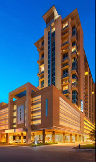 A tall, modern hotel building with multiple balconies and illuminated windows against a twilight sky. The street in front is quiet and the building displays the number 5250.