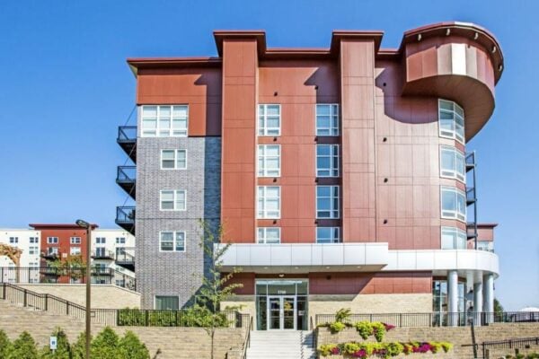 Modern four-story apartment building with red and gray exterior, balconies, large windows, and landscaped entryway under a clear blue sky.