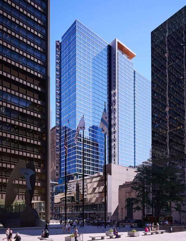 A modern glass office building stands between two older dark skyscrapers, with flags flying and people walking in a plaza below on a clear day.