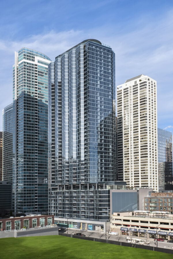 A group of modern high-rise buildings with glass exteriors, set against a blue sky, with a green lawn and city streets in the foreground.