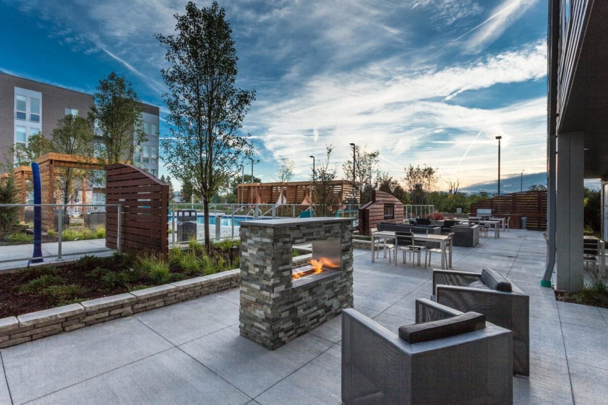 Modern outdoor patio area with a stone fireplace, seating, dining tables, and landscaping, set against a backdrop of apartments and a blue sky with clouds.