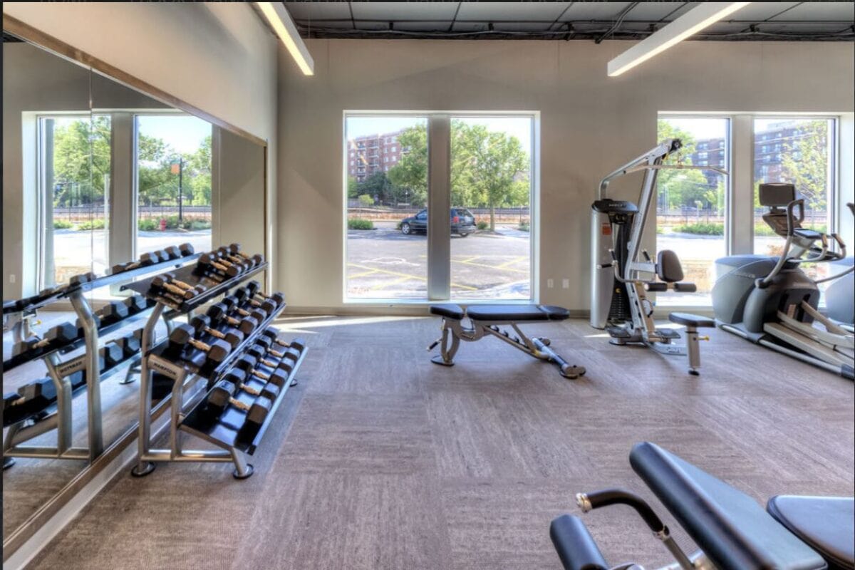 A gym with free weights, benches, a weight machine, and cardio equipment, featuring large windows with a view of a parking lot and buildings outside.