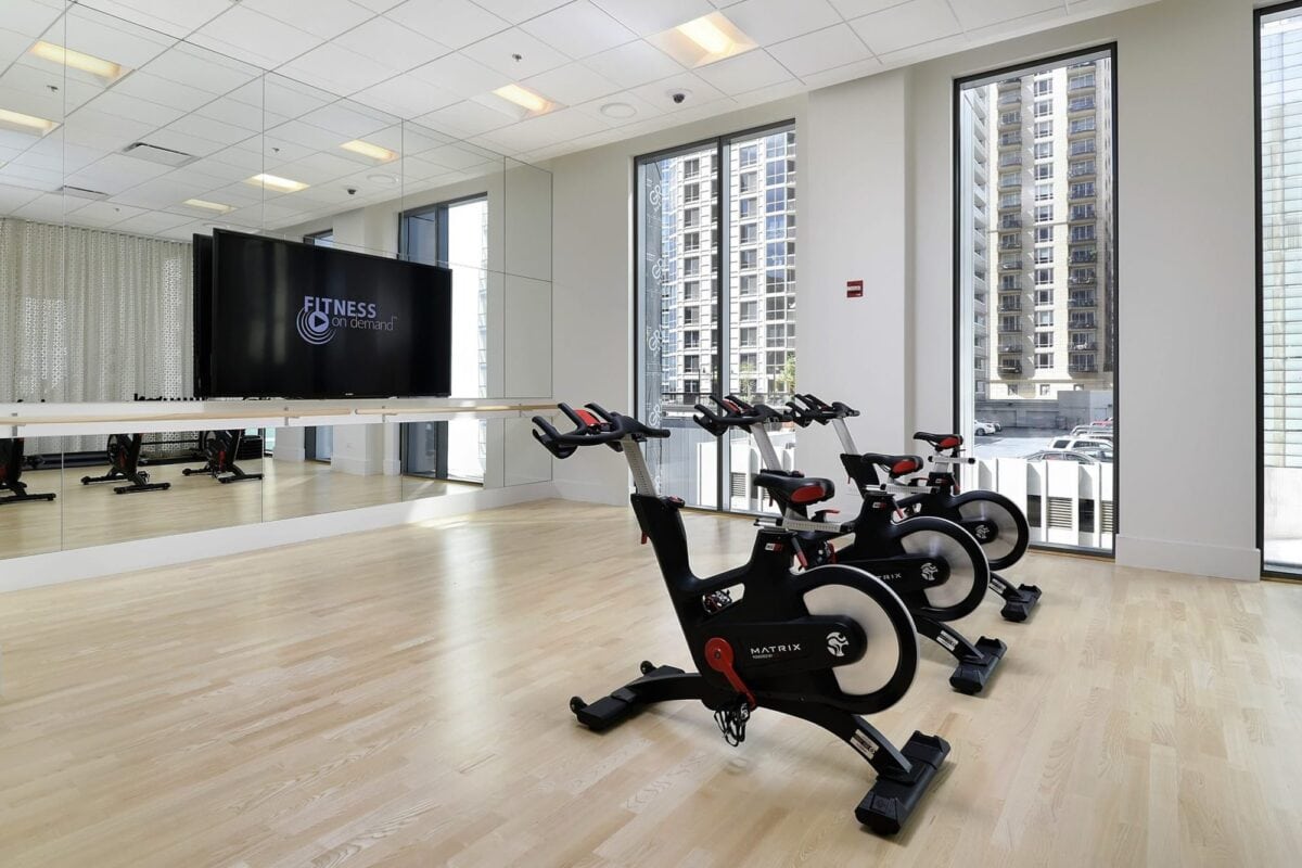 Three stationary exercise bikes are lined up in a bright gym room with large windows, a wall mirror, and a TV displaying “Fitness Center.”.