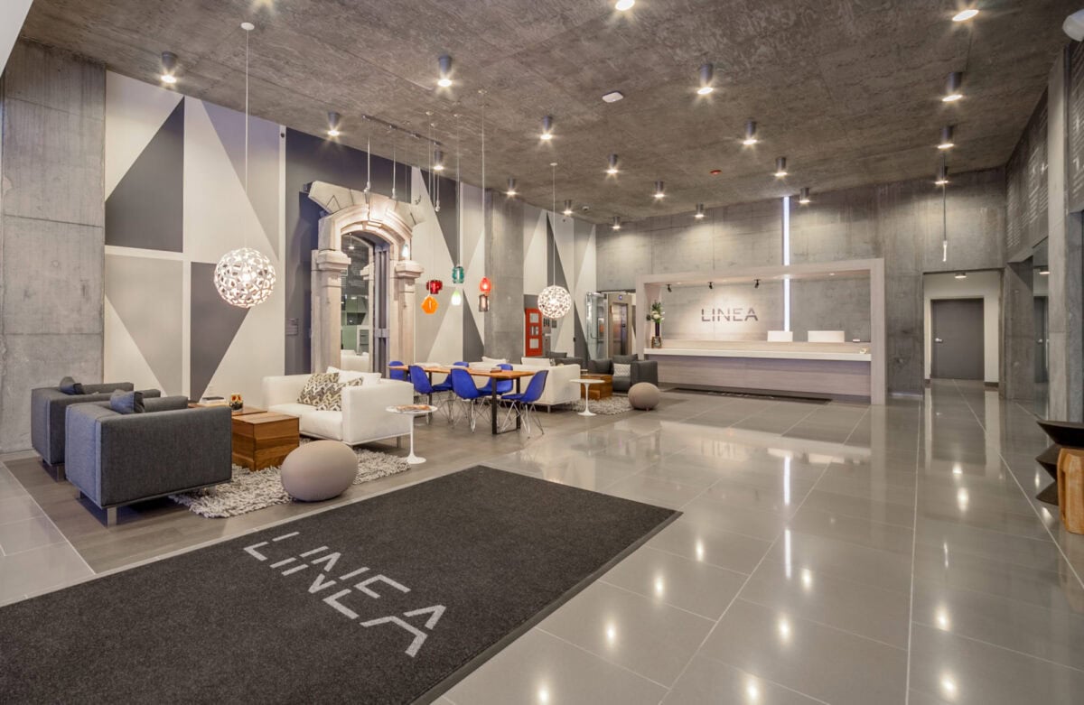 Modern lobby with gray tile floors, geometric wall art, contemporary furniture, spherical pendant lights, and a reception desk labeled "LINEA.