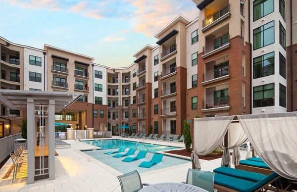Modern apartment complex courtyard featuring a swimming pool with lounge chairs, cabanas, and multiple-story buildings surrounding the pool area.
