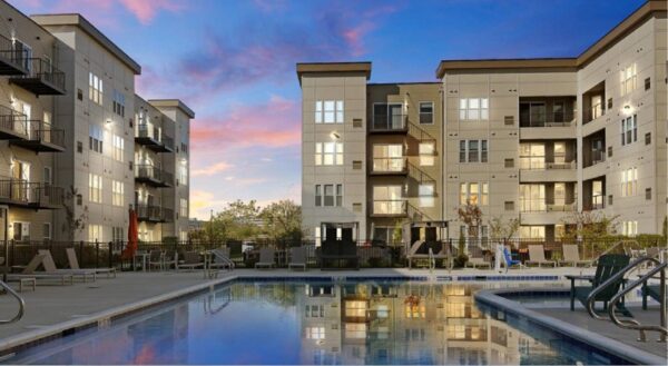 Modern apartment buildings at sunset with illuminated windows, overlooking an outdoor swimming pool and lounge chair area.