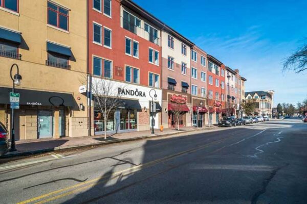 Street view of multi-story buildings with various storefronts, including a Pandora store, under a clear blue sky with minimal traffic and empty sidewalks.