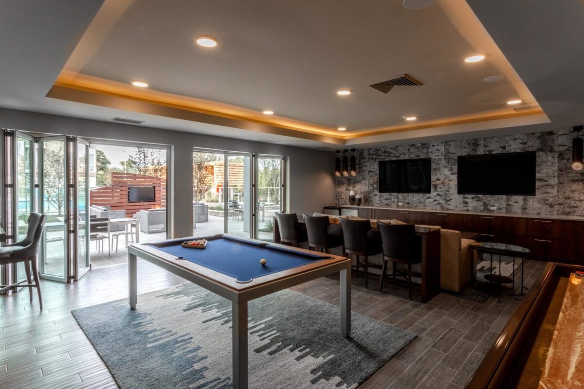 Modern game room with pool table, lounge seating, TV screens, and sliding glass doors opening to an outdoor patio area.