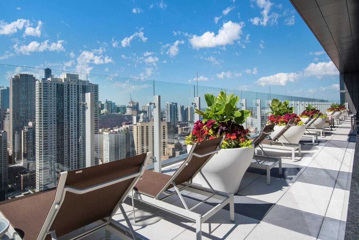 A rooftop terrace with lounge chairs and large planters overlooks a city skyline with tall buildings under a blue sky with scattered clouds.