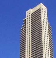 A tall, modern high-rise apartment building with balconies, set against a clear blue sky.