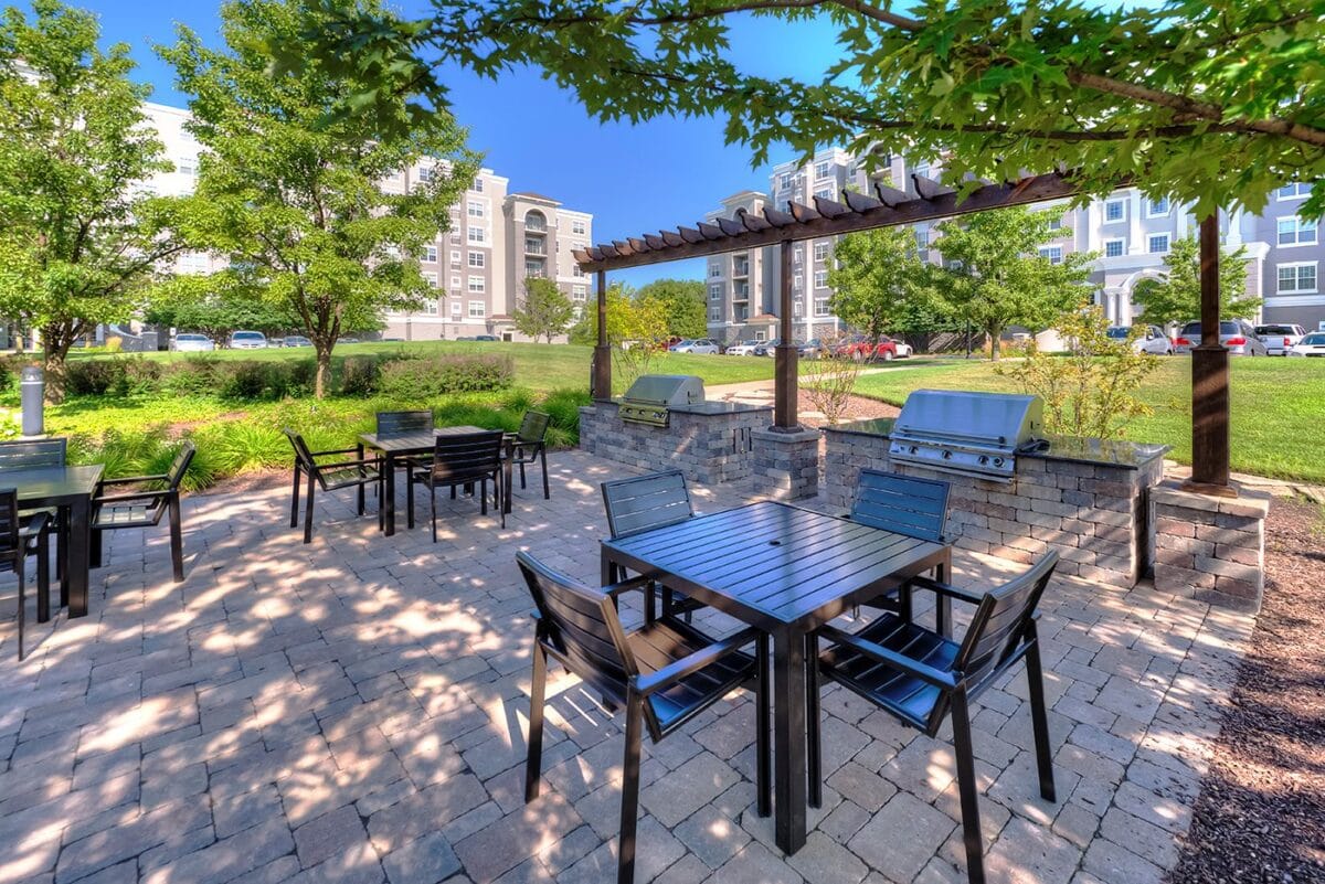 Outdoor patio with black tables and chairs, shaded by trees, featuring built-in grills under a wooden pergola, with apartment buildings visible in the background.