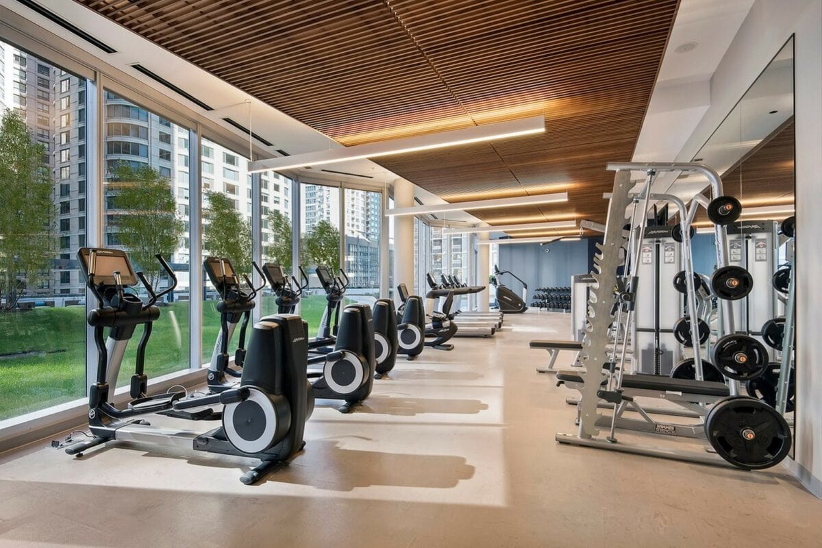 Modern gym with treadmills, elliptical machines, and weight equipment next to large windows overlooking buildings and greenery. Wooden ceiling and mirrors add to the contemporary design.
