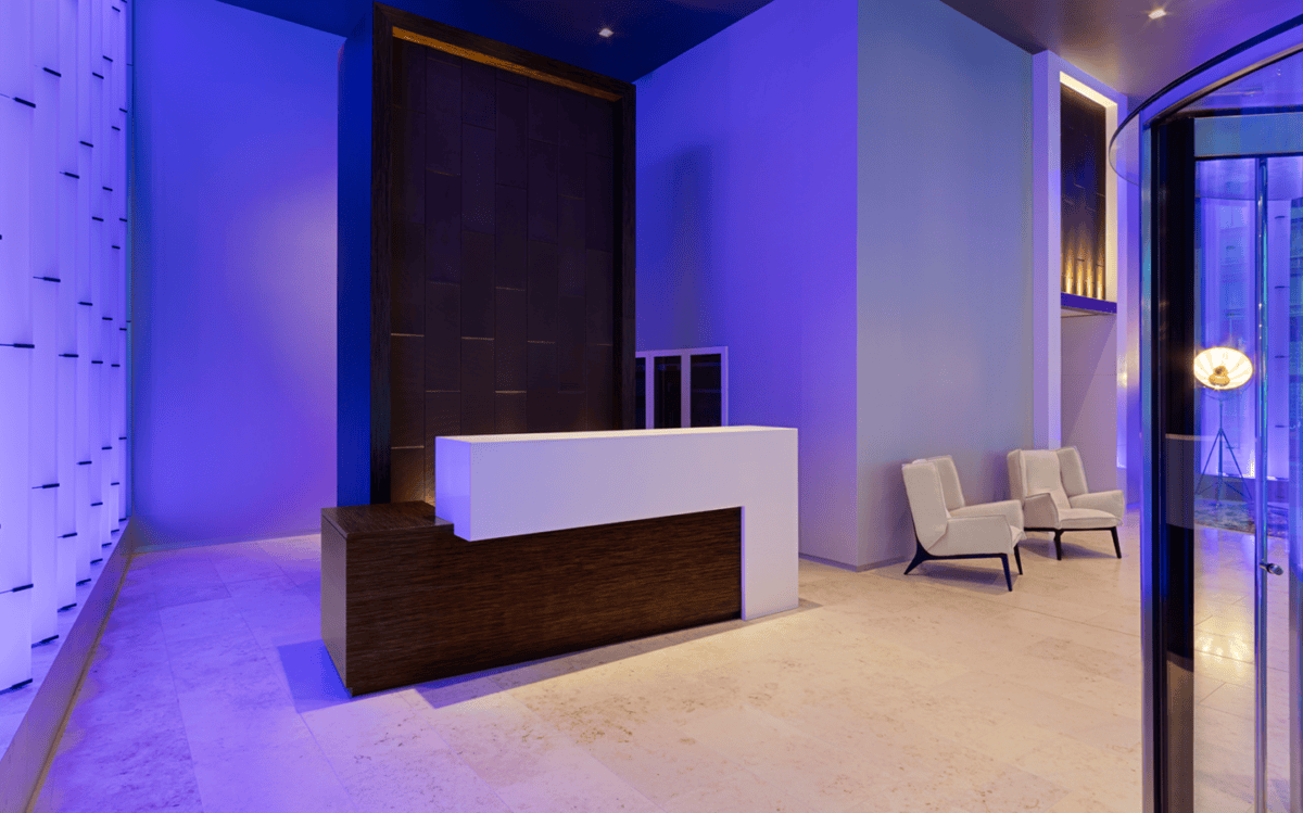 Modern lobby with a white and dark wood reception desk, two beige chairs, light-colored floor, blue accent lighting, and large window to the right.