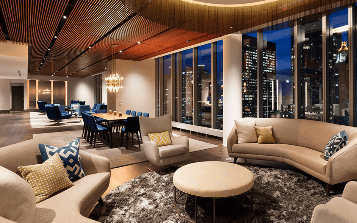 A modern lounge with curved sofas, armchairs, a round ottoman, and a dining area, featuring floor-to-ceiling windows overlooking a city skyline at night.