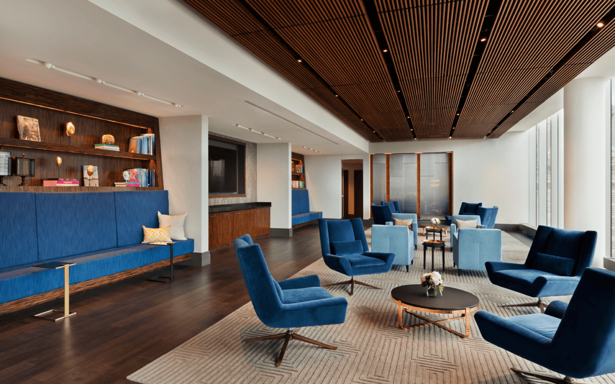 Modern lounge with blue chairs, built-in seating, bookshelves, round tables, large windows, and wood-paneled ceiling.