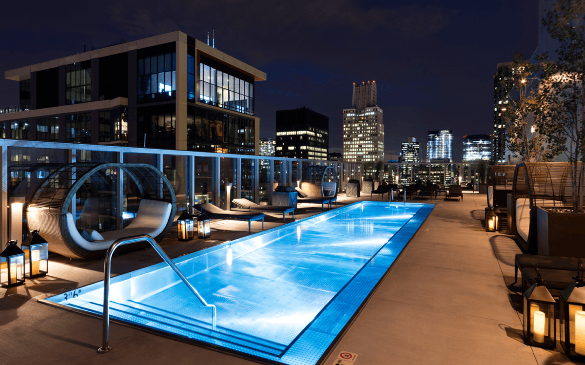 Rooftop swimming pool at night with modern lounge chairs, hanging pods, and city buildings illuminated in the background.