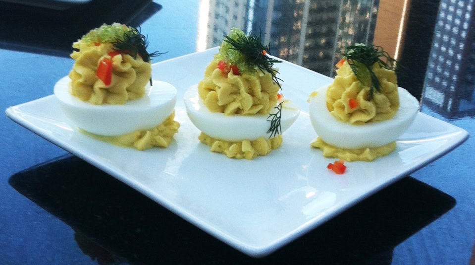 Three deviled eggs topped with herbs and small diced red garnish are arranged on a white square plate, set against a cityscape background.