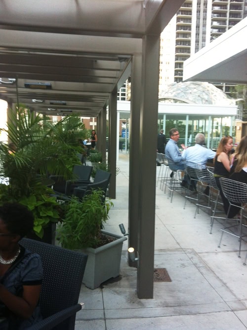 Outdoor restaurant patio with people seated at tables, surrounded by potted plants and city buildings in the background.
