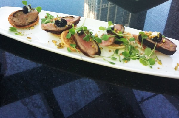 A white rectangular plate with sliced meat on toasted rounds, topped with microgreens and a dark garnish, set on a reflective glass surface.