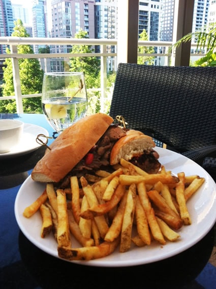 A plate of French fries and a sandwich is served on an outdoor table with a glass of water, city buildings, and greenery visible in the background.