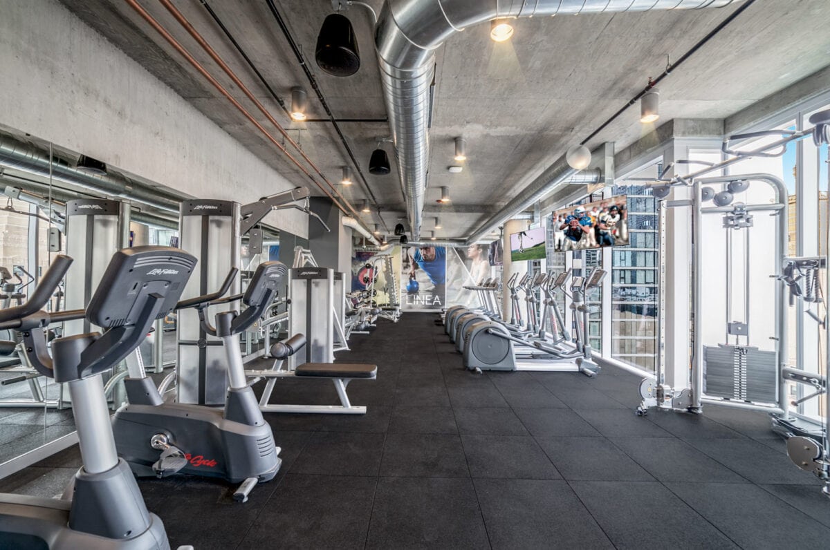 Modern gym with various cardio machines, weight equipment, large windows, and mirrors along the walls under exposed piping and ceiling.