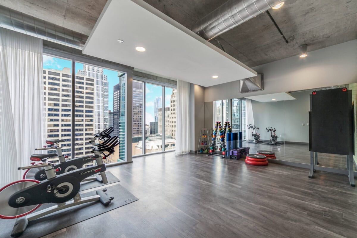 Modern gym with exercise bikes, free weights, fitness equipment, large windows with city views, and a mirrored wall.