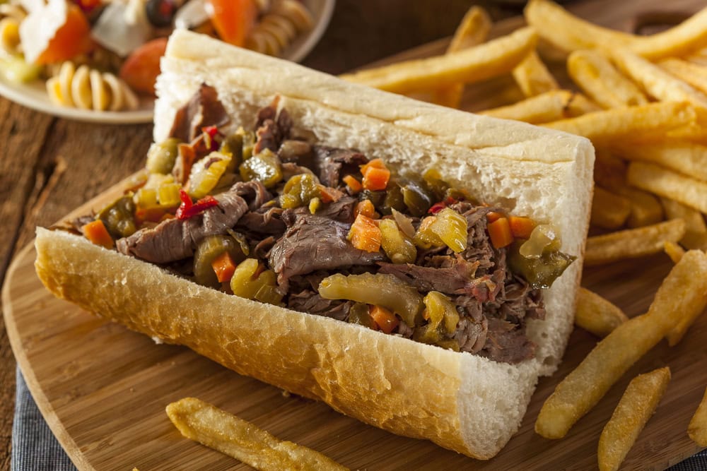 Italian beef sandwich topped with chopped pickled vegetables on a wooden tray, served with a side of French fries.