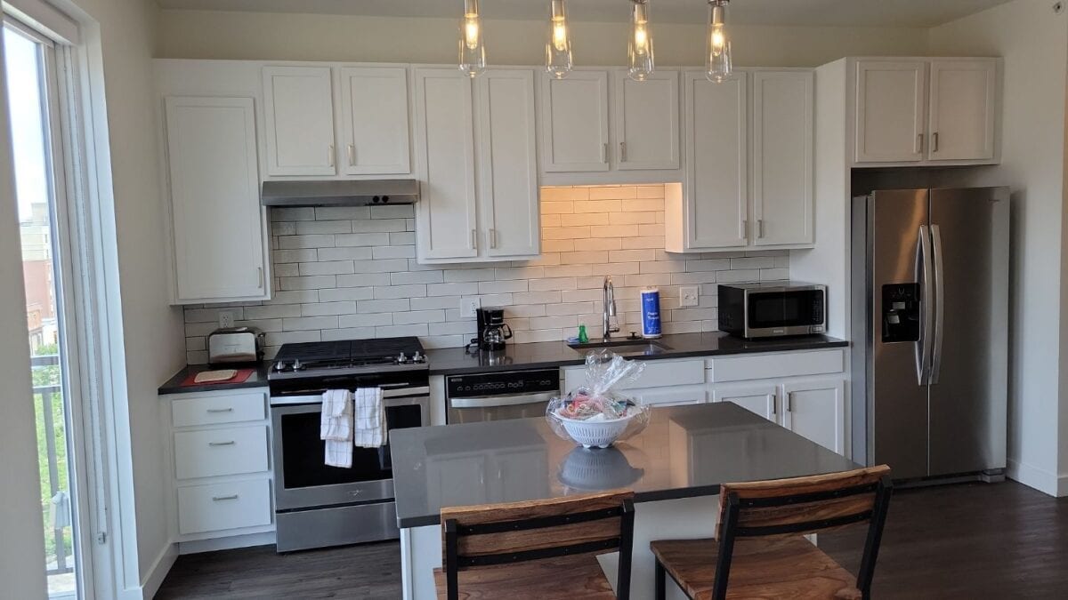 Modern kitchen with white cabinets, stainless steel appliances, subway tile backsplash, an island with two wooden chairs, and a bowl on the counter under hanging lights.
