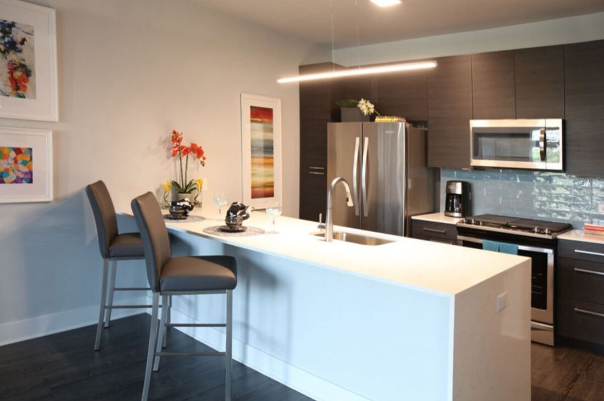 Modern kitchen with dark wood cabinets, stainless steel appliances, a white island with two bar stools, and colorful artwork on the walls.