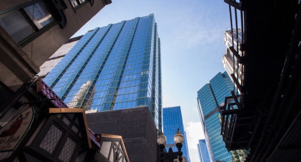 Tall modern glass buildings rise against a blue sky, framed by older structures and a streetlamp in the foreground.