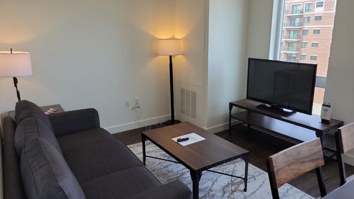 A modern living room with a gray sofa, two lamps, a coffee table with papers and a remote, a TV on a stand, two chairs, and a window showing an apartment building outside.
