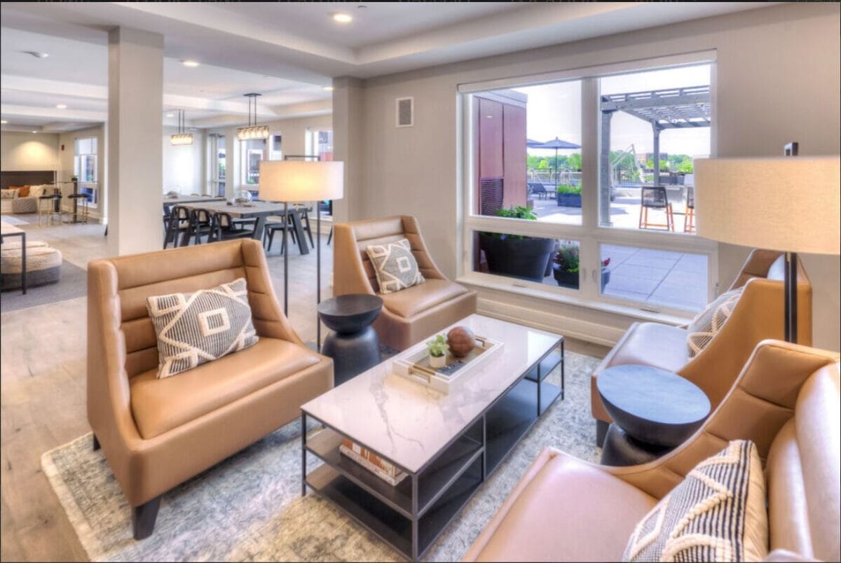 Modern living room with tan leather chairs, glass coffee table, floor lamps, patterned pillows, and large window showing an outdoor patio. Dining area visible in the background.