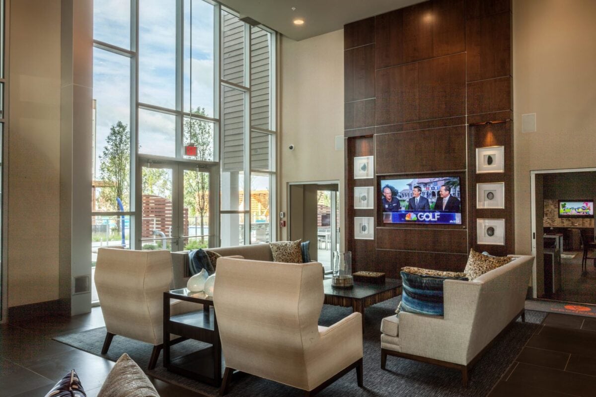 Modern lounge area with cream-colored chairs, a coffee table, large windows, and a wall-mounted TV displaying a golf program. Natural light fills the room.