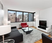 Modern living room with large windows, city view, black sofa, red chair, glass coffee table, TV, and white walls with simple decor.