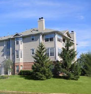 Three-story apartment building with beige siding, brick detailing, and multiple chimneys, surrounded by green grass, trees, and a clear blue sky.