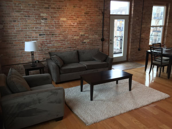 Living room with exposed brick walls, two gray sofas, a coffee table on a white rug, a lamp, and a dining table by large windows.