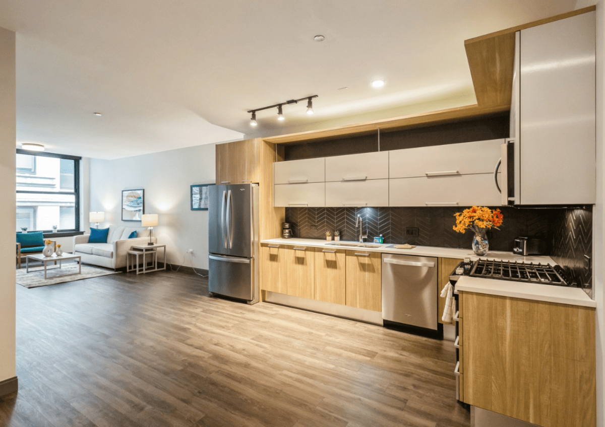 Modern open-plan kitchen with stainless steel appliances, wood cabinets, and a vase of flowers on the counter, adjoining a living area with a large window and sofa.