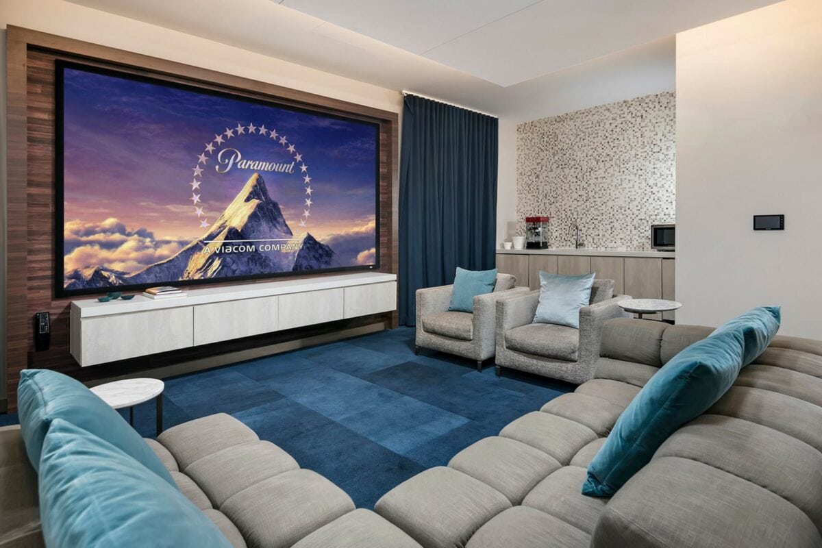 A modern home theater room with a large movie screen, comfortable seating, blue accents, and a small kitchenette in the background.