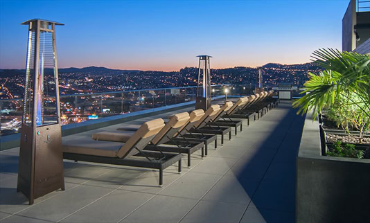 A modern rooftop patio with lounge chairs, outdoor heaters, potted plants, and a cityscape view at dusk.