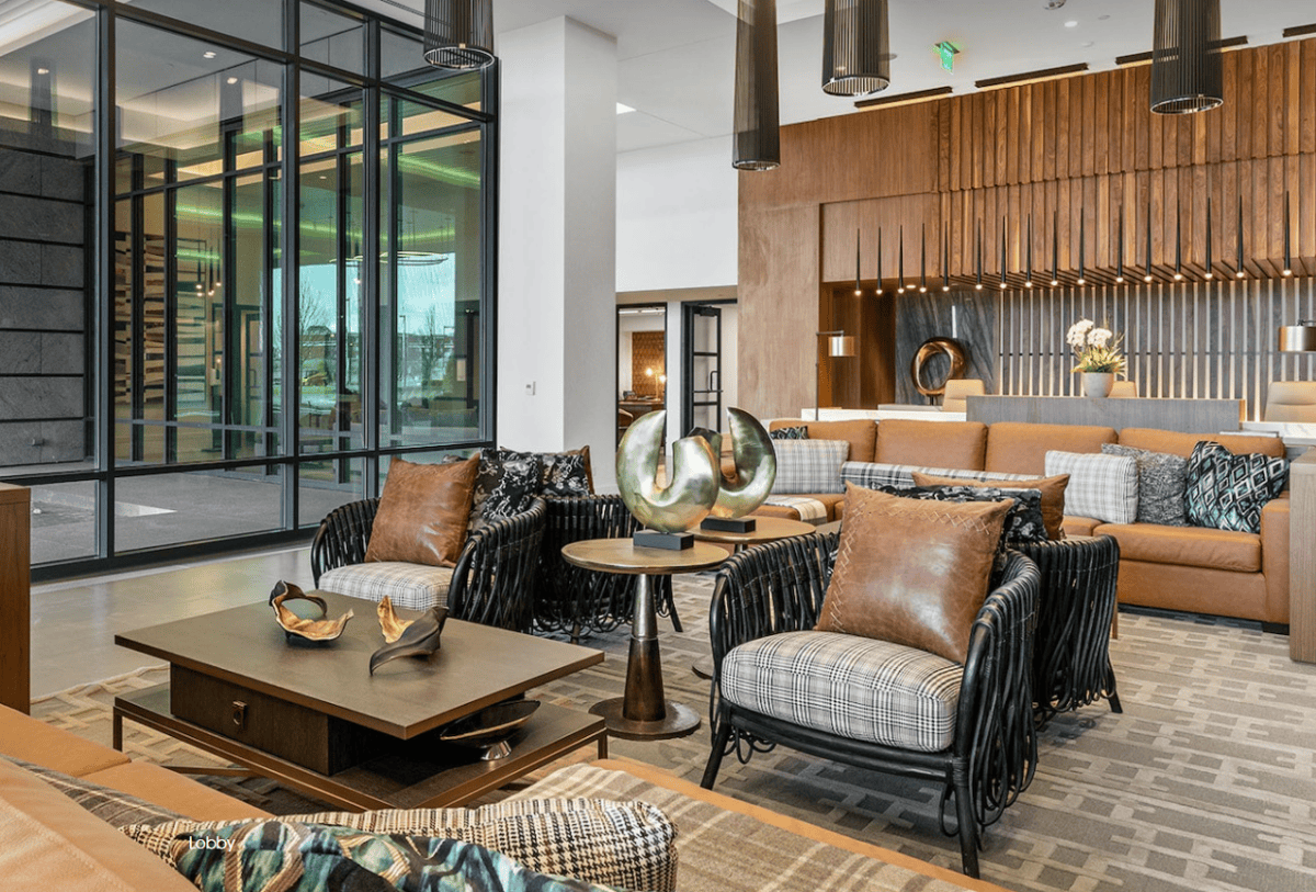 Modern hotel lobby with contemporary furniture, decorative pillows, metal sculptures on tables, and wood paneling on the back wall. Large windows provide natural light.